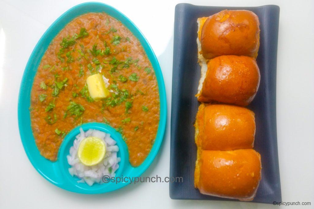 butter toasted pav is served with bhaji in a oval shaped green plate served with butter cube and fresh coriander on its top and chopped onion & lemon wedge on a side of the plate & butter toasted pav is served on a black rectangular plate kept side by side on a white table top